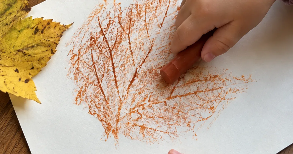 A child rubbing a crayon over paper to reveal the shape of a leaf
